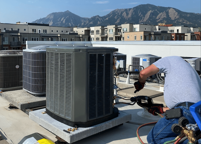 Technician repairing rooftop HVAC unit with cityscape and mountains in the background.